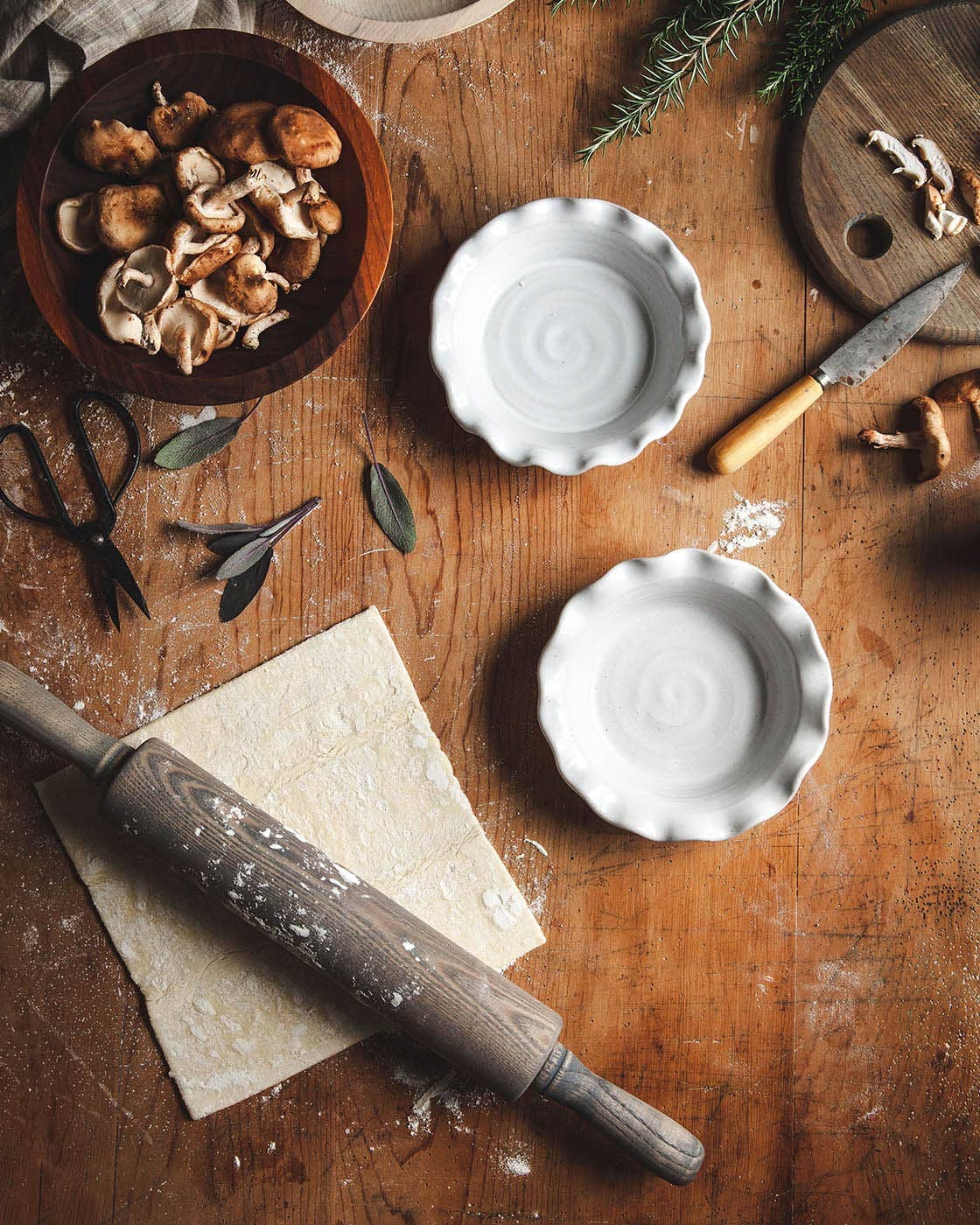 Wooden table with ingredients and kitchen tools including a rolling pin, bowls, and herbs.