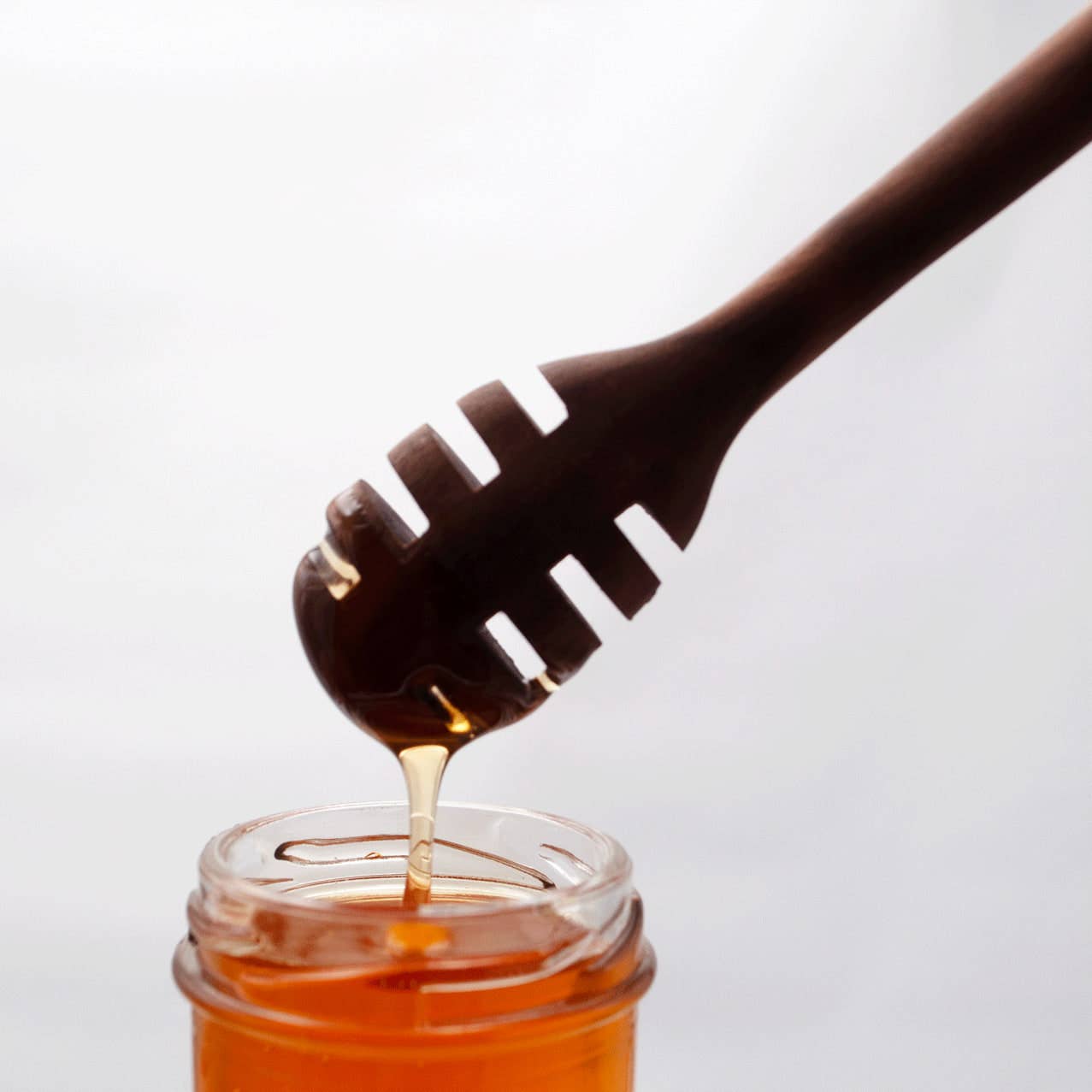 Wooden honey dipper with honey dripping into a jar on a white background