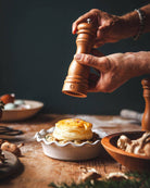 Hand using a pepper grinder over a dish of food on a wooden table.